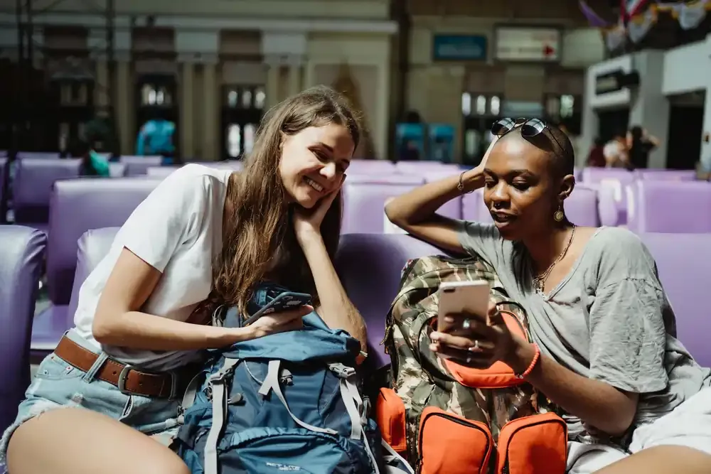 Two female travelers sitting in an airport or train station lounge, smiling and looking at a smartphone together, with backpacks beside them.