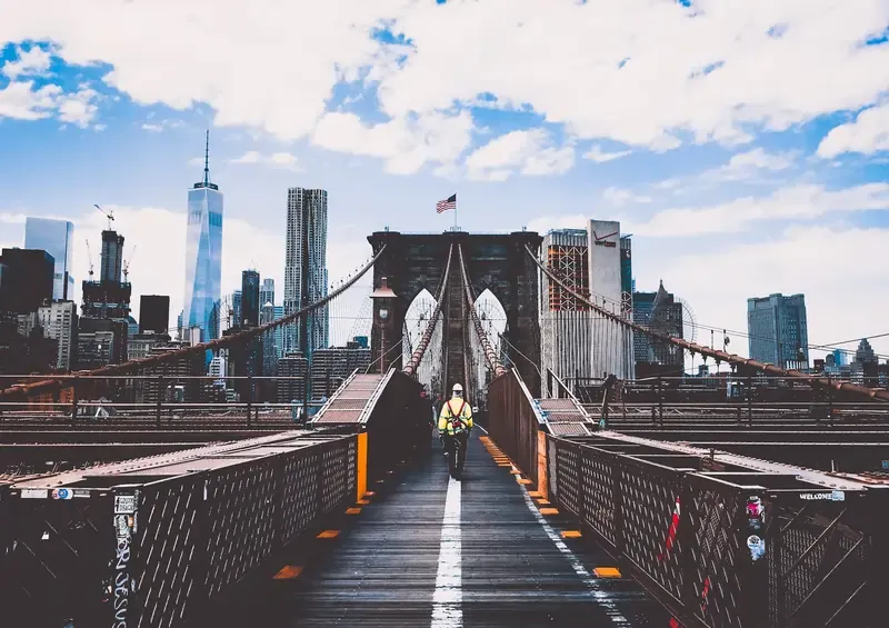 Worker in high-visibility vest walking on the Brooklyn Bridge with the New York City skyline in the background.