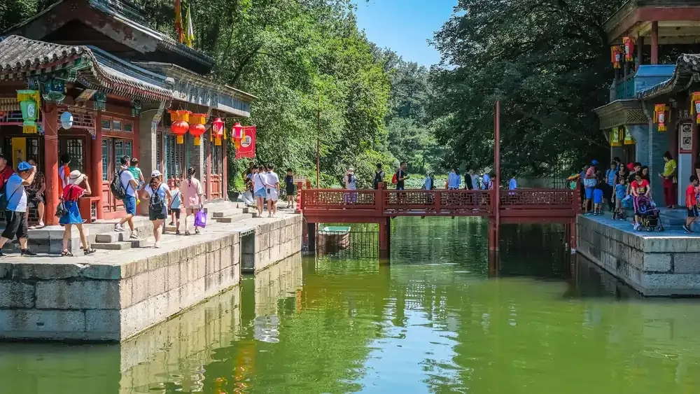 Visitors walking along traditional Chinese buildings and crossing a red bridge over a canal at the Summer Palace in Beijing.