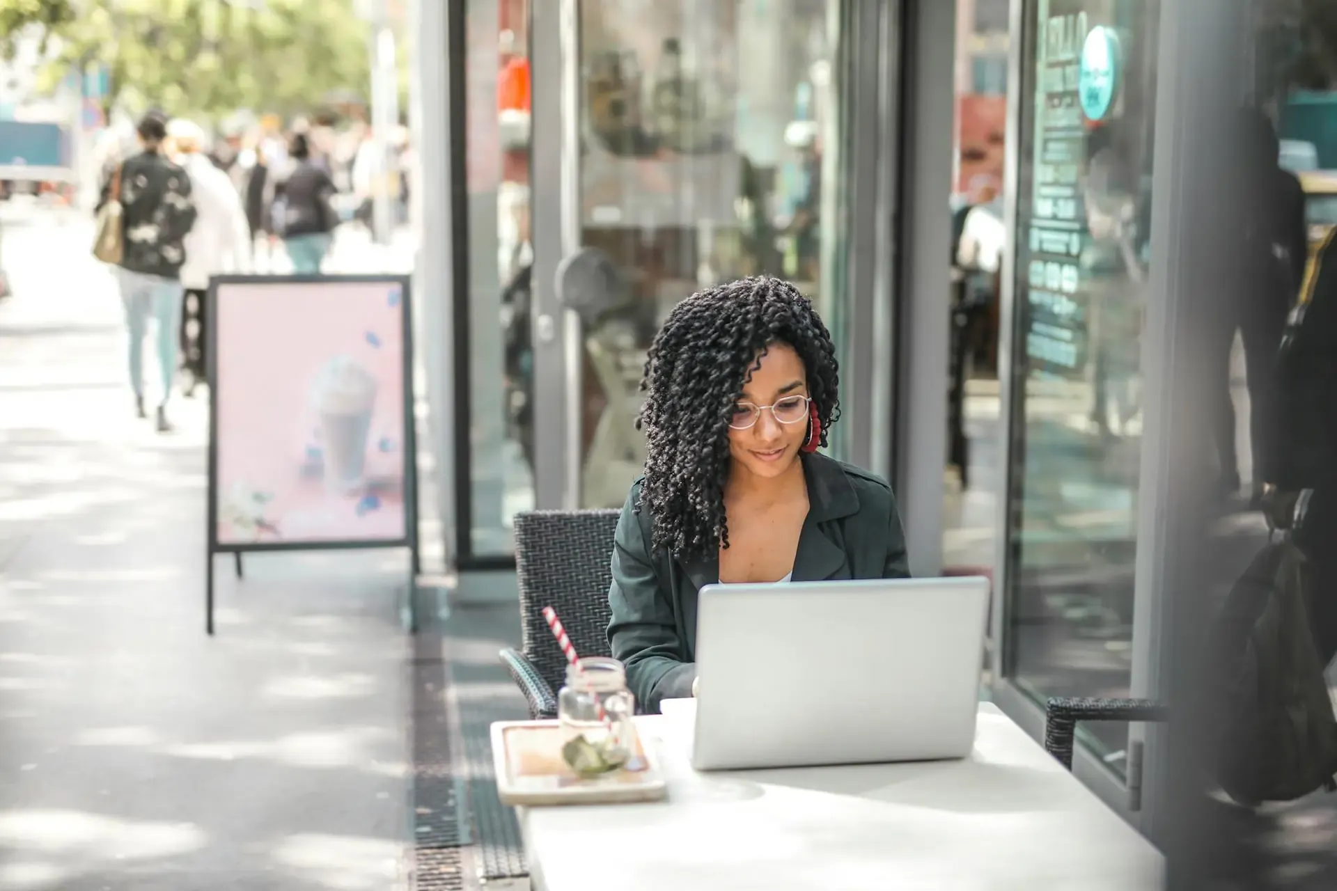 Smiling woman working on a laptop at an outdoor café table, with a drink beside her. 