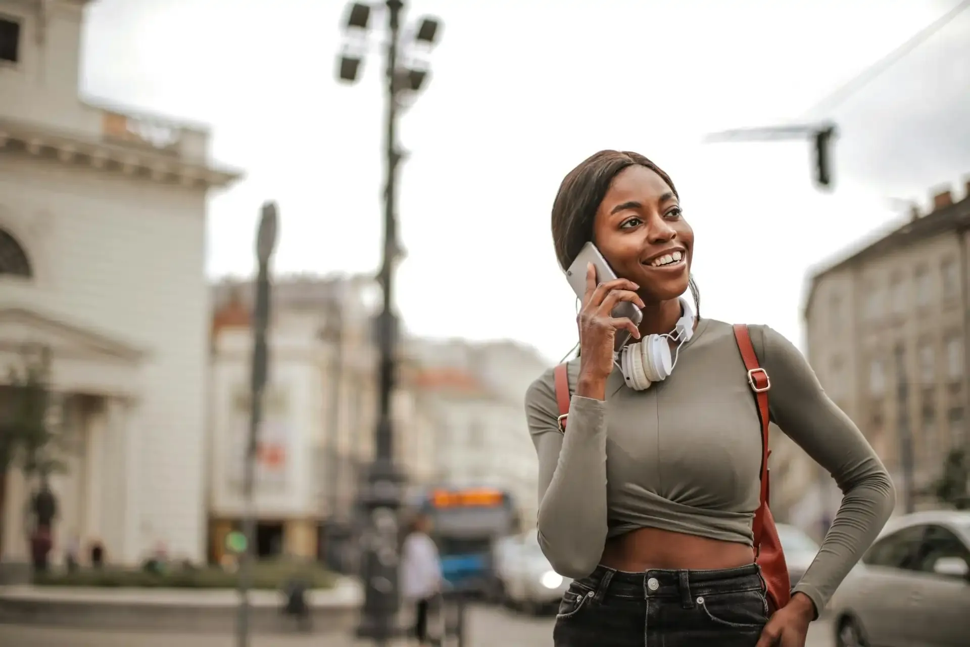 Smiling woman with headphones and backpack using her phone outdoors.