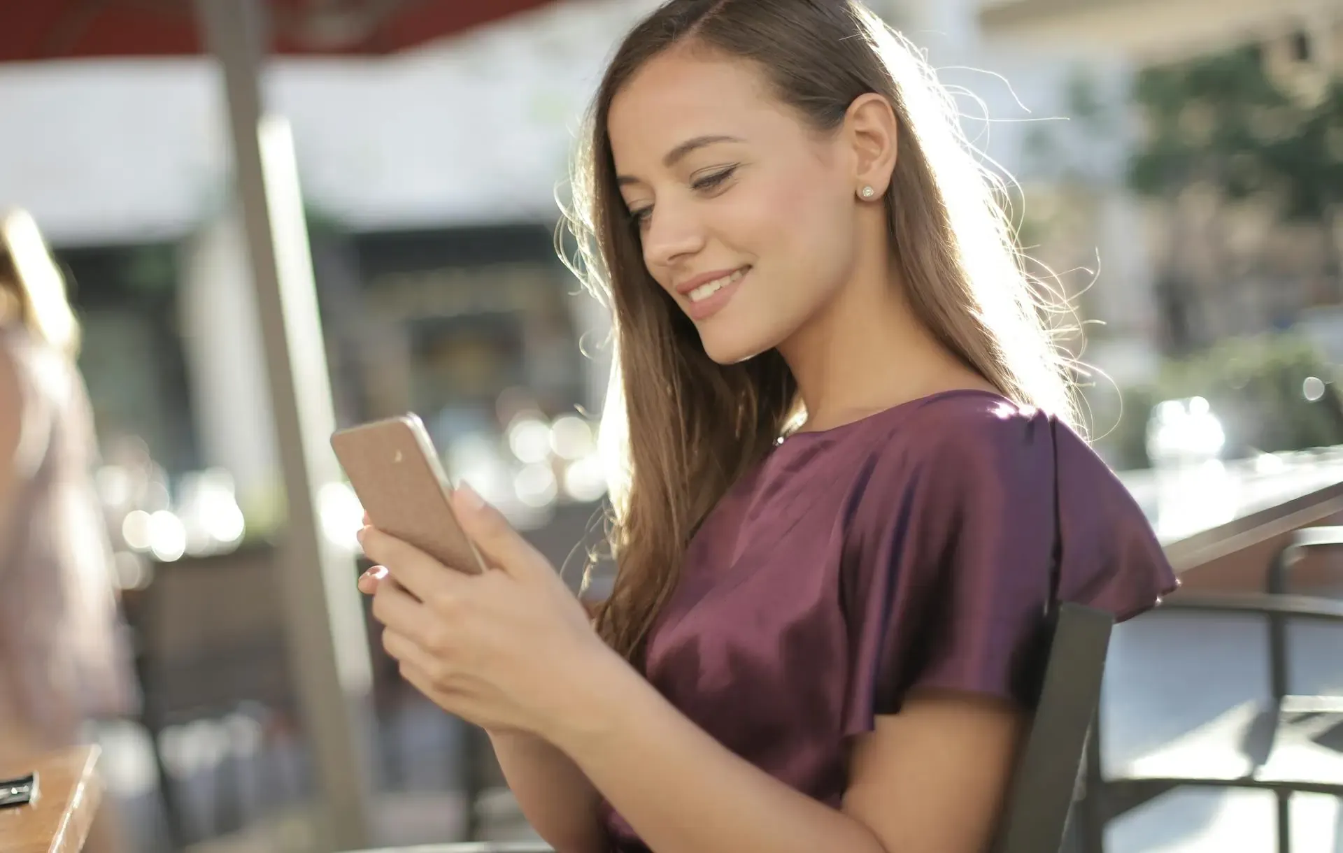 Smiling woman sitting outdoors, using her smartphone, with sunlight highlighting her hair and a blurred city background.
