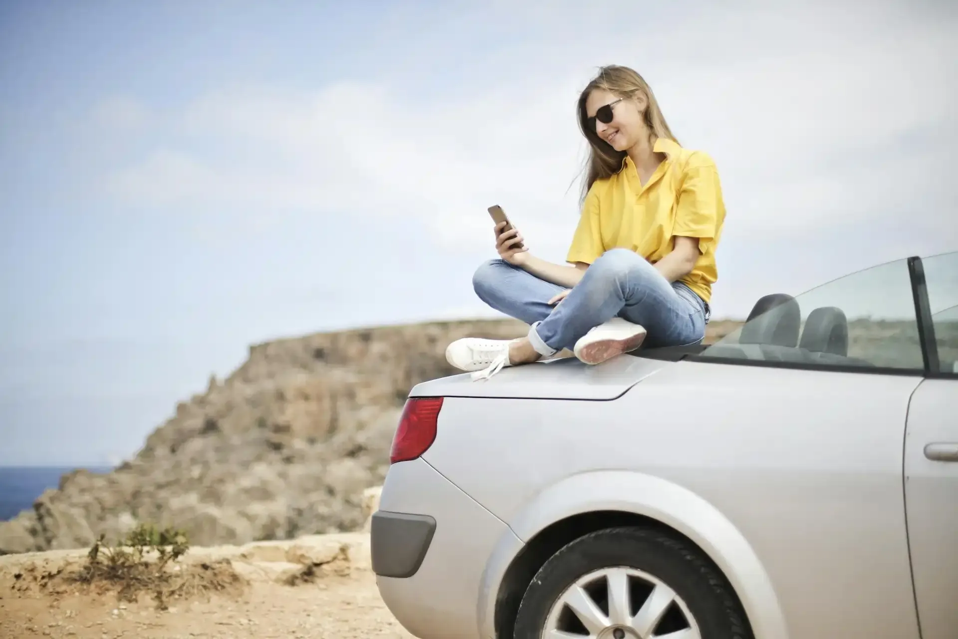 Woman sitting on a convertible car by the coast, using a smartphone during travel.