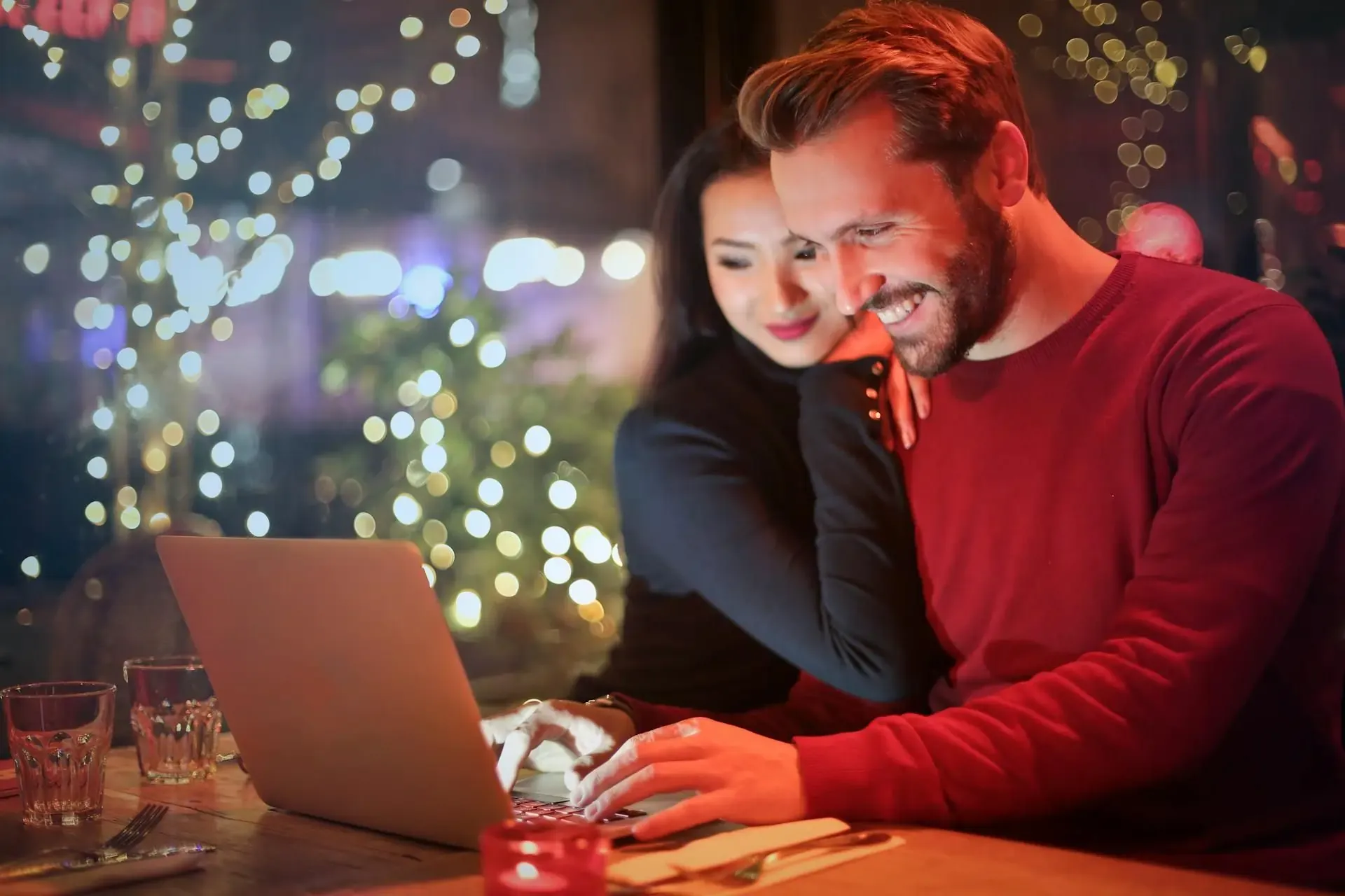 Smiling couple using a laptop in a cozy restaurant setting, with festive lights in the background