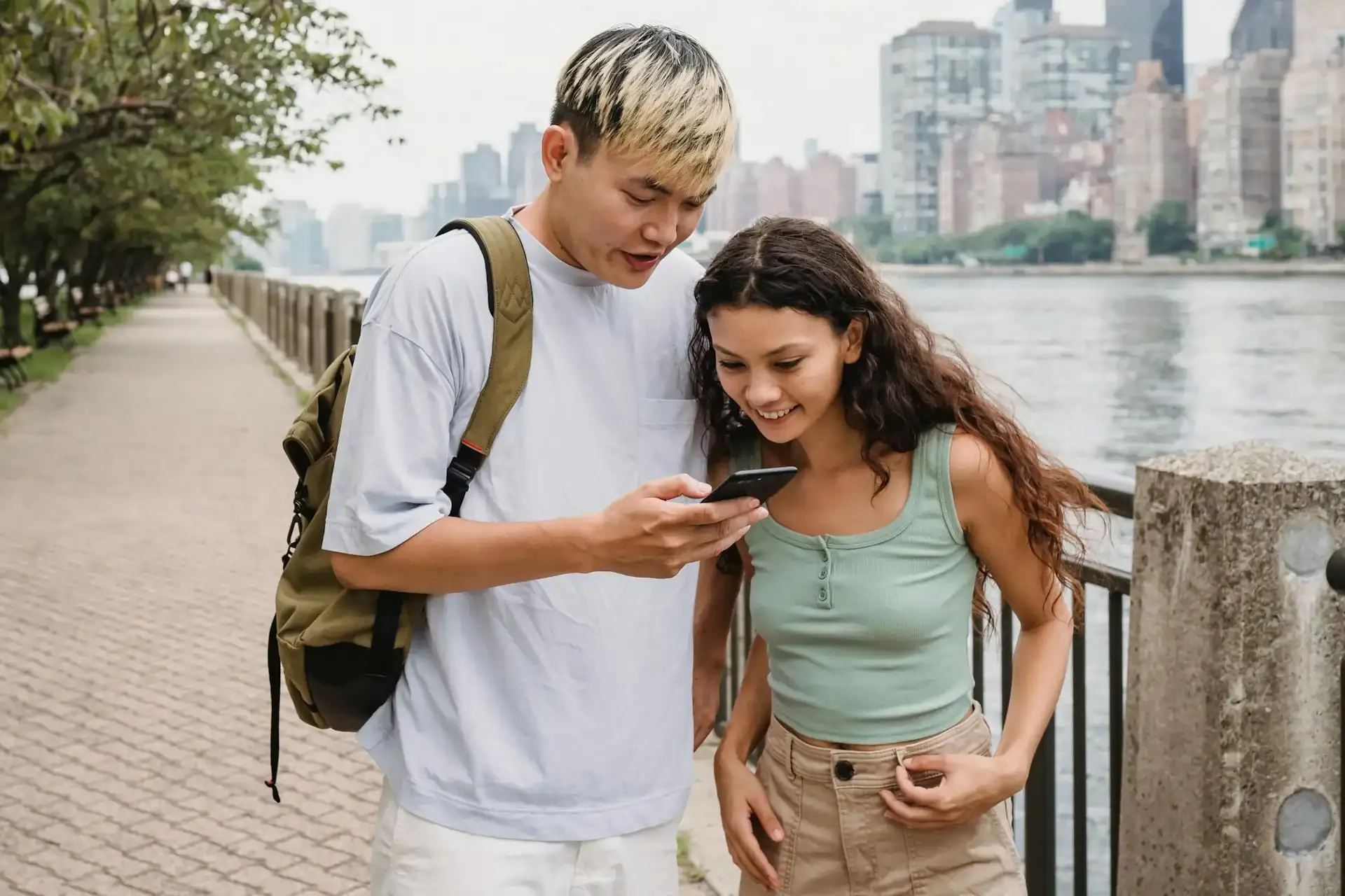 Two young travelers looking at a smartphone while walking by a riverside in the city