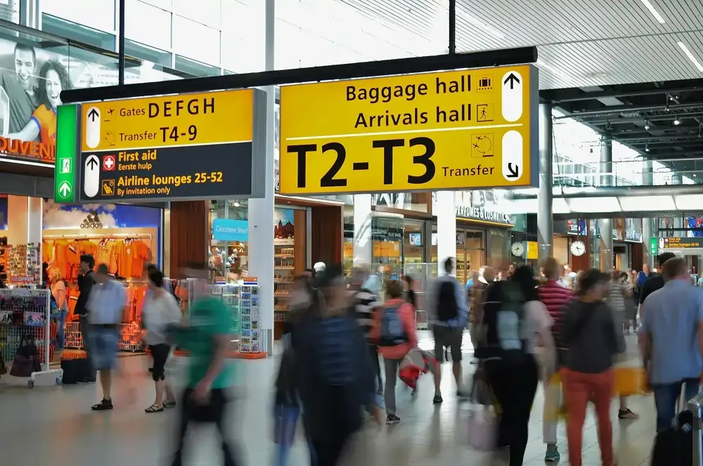 Busy airport terminal with travelers walking under yellow directional signs for transfers and arrivals