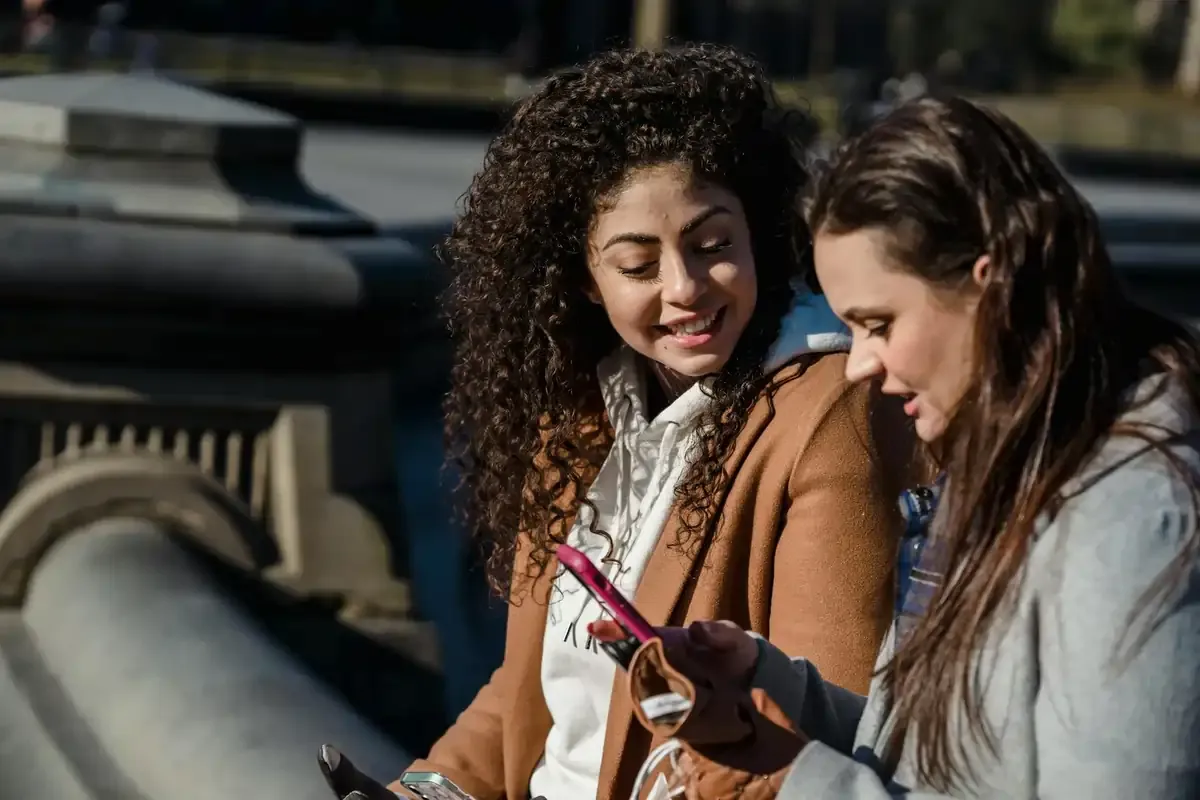 Two women on an outdoor bench looking at a smartphone together, illustrating sharing an eSIM data plan for travel connectivity.