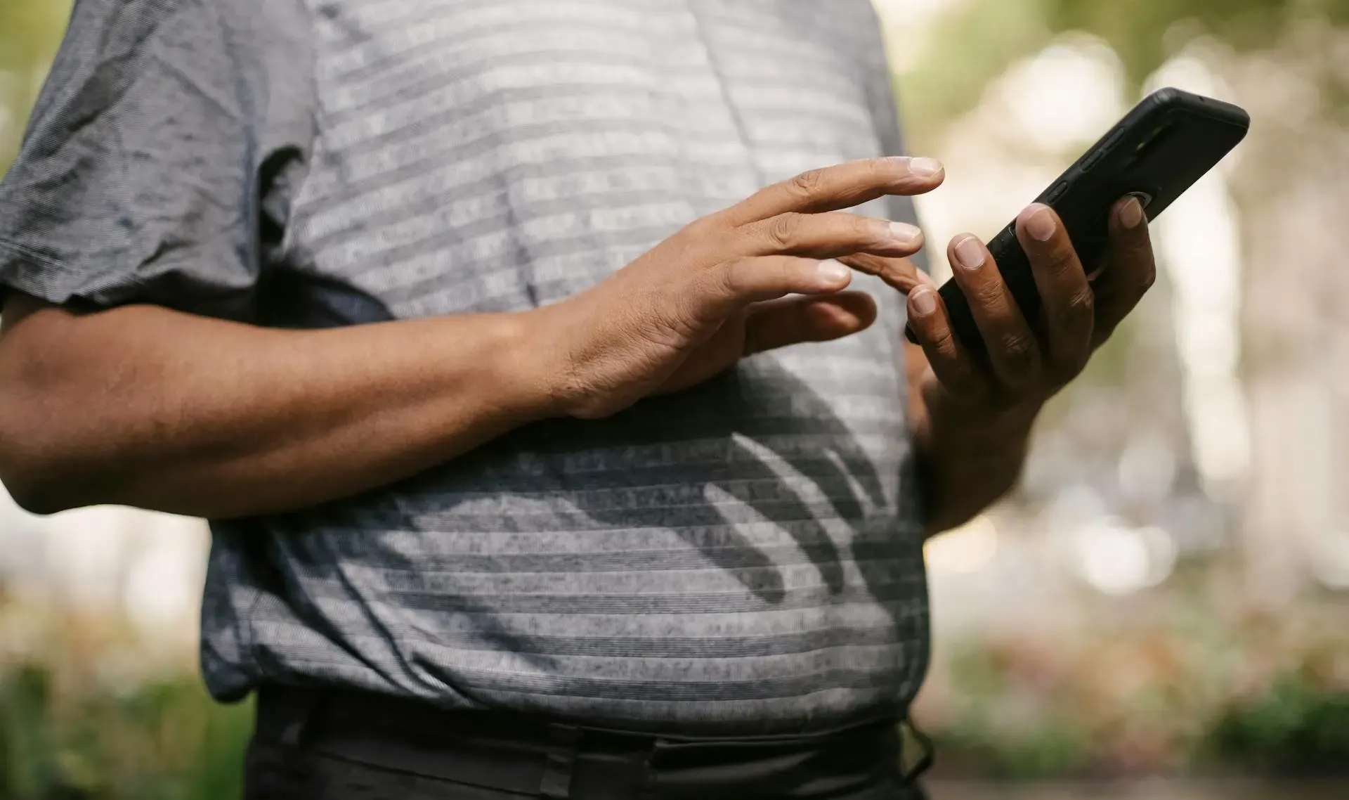 Close-up of a man using a smartphone outdoors, with one hand holding the phone and the other tapping the screen, wearing a striped grey shirt.