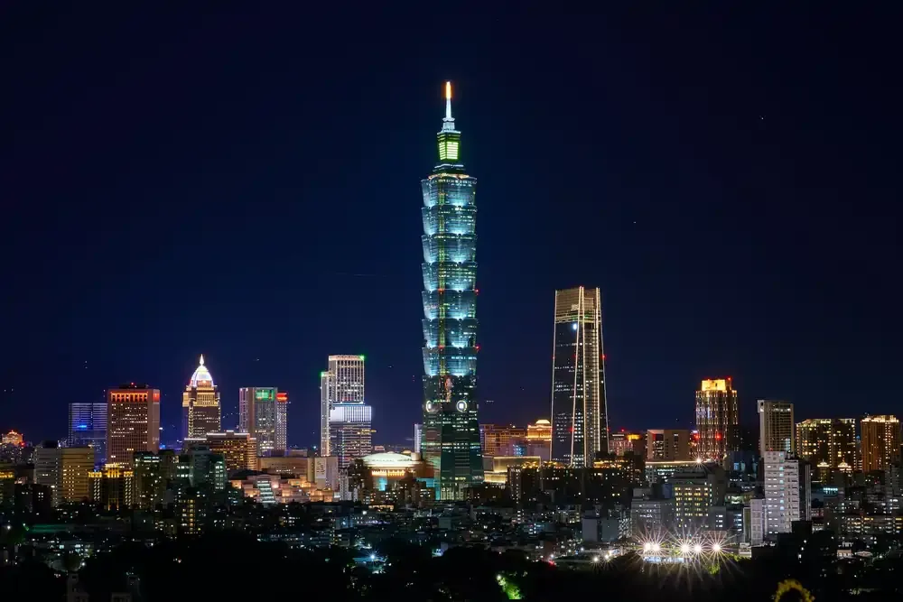 Nighttime cityscape of Taipei, Taiwan, with the brightly lit Taipei 101 skyscraper standing prominently in the center among other illuminated buildings.