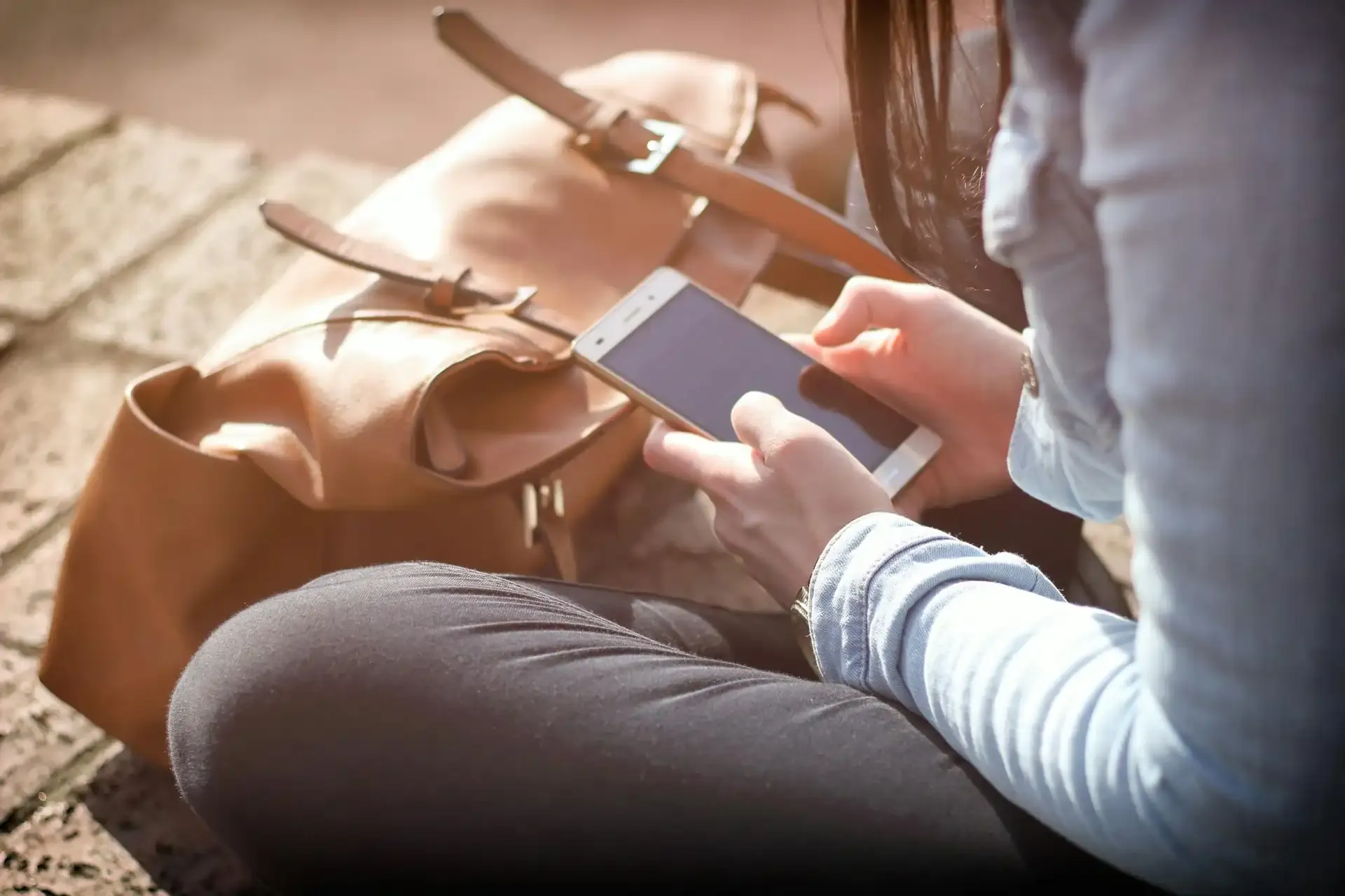Person sitting outdoors using a smartphone next to a tan leather bag, checking messages or settings on their mobile device.