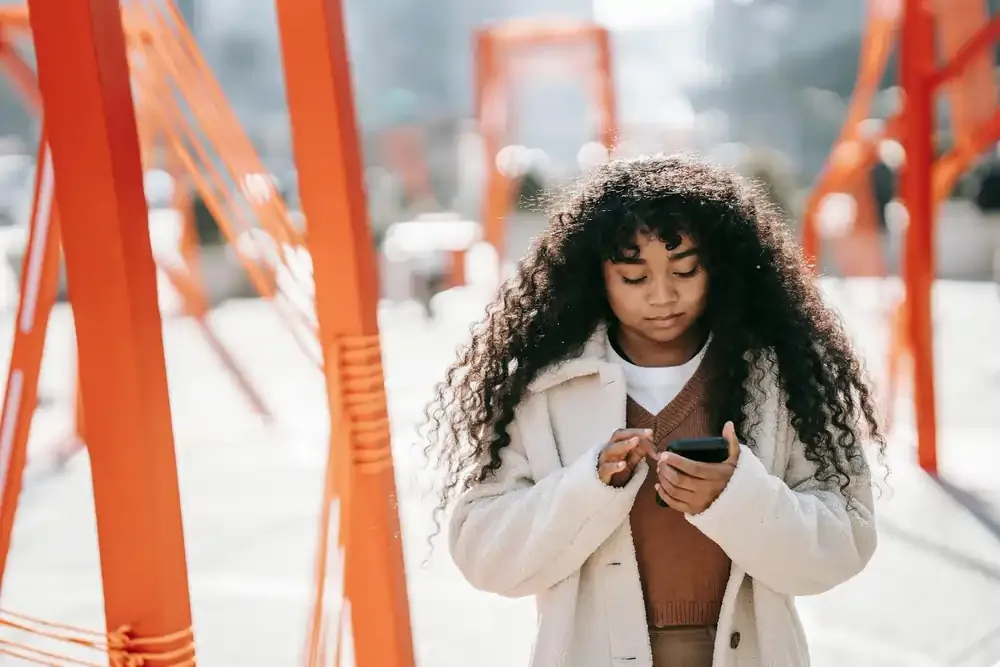Young woman with curly hair using a smartphone while walking through a public art installation with bright orange beams.