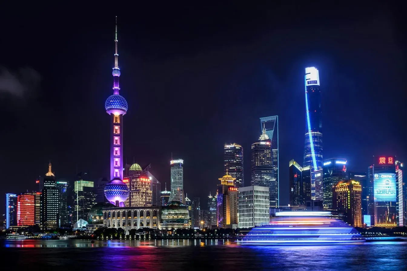 Night view of the illuminated Shanghai skyline featuring the iconic Oriental Pearl Tower, Shanghai Tower, and modern skyscrapers reflecting on the Huangpu River.