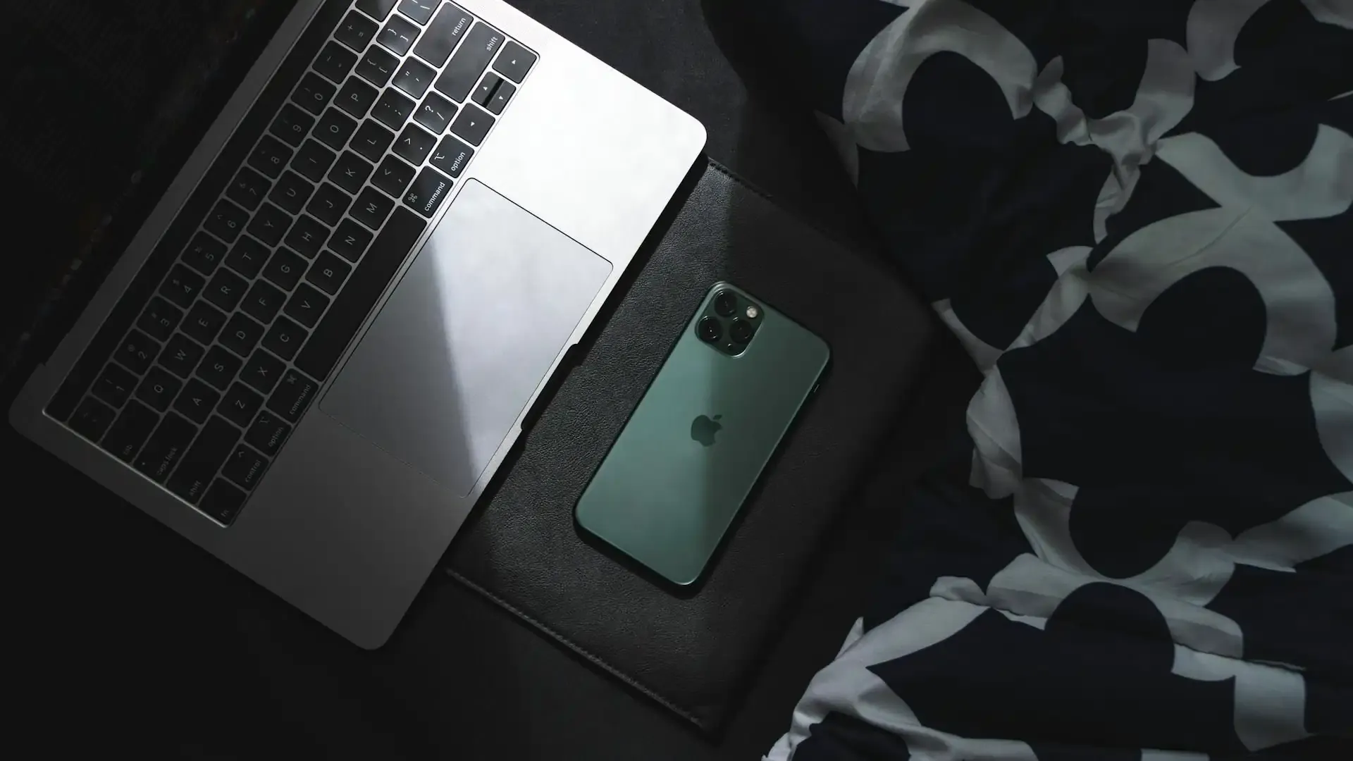 iPhone and MacBook placed beside each other on a dark bedspread, illuminated by a single light source, symbolizing minimal tech setup and mobile connectivity.