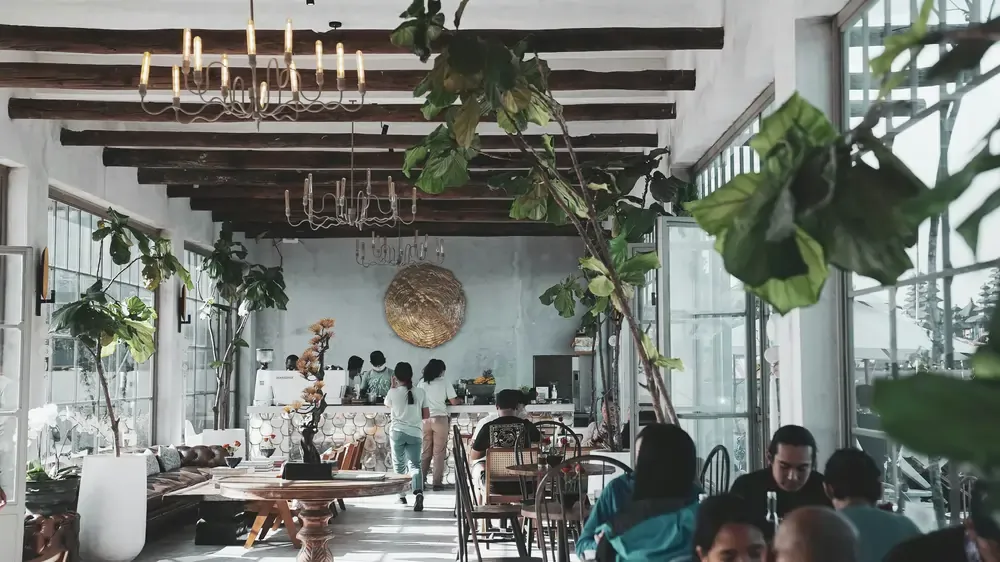 Bright café interior with high ceilings, wooden beams, indoor plants, and people seated at tables in a relaxed setting.