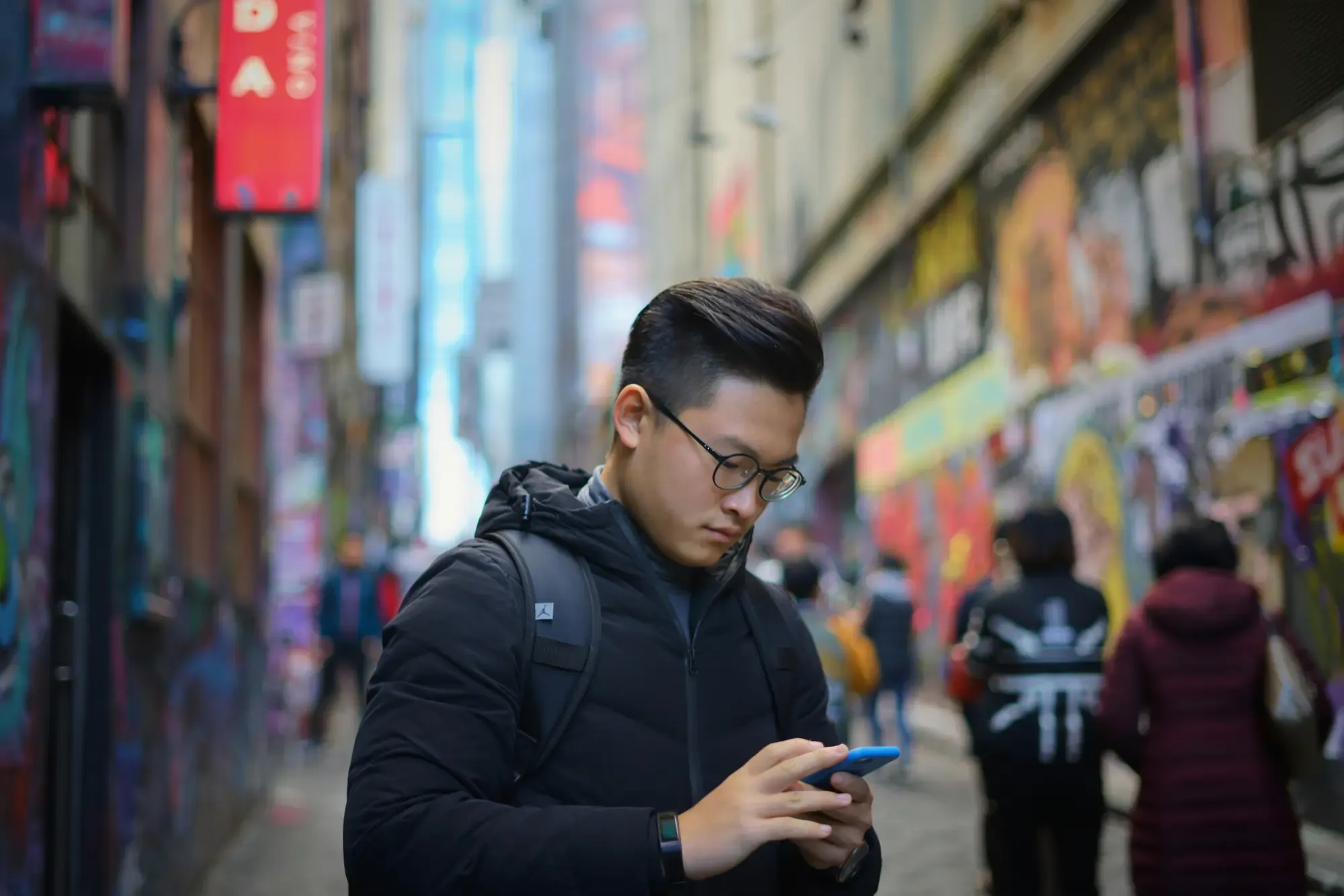 Traveler wearing glasses and a black jacket using a smartphone while standing in a vibrant city laneway lined with colorful street art and storefronts.