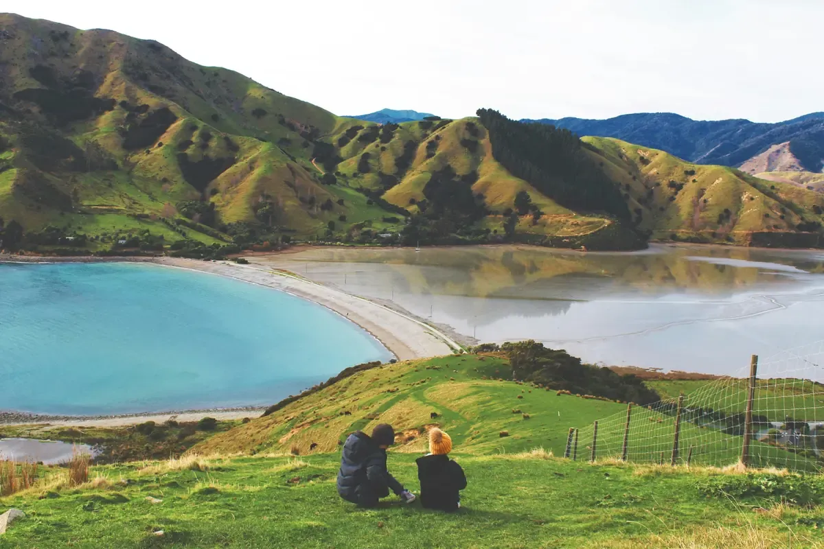 Two people overlooking a turquoise coastal bay surrounded by green hills in New Zealand.