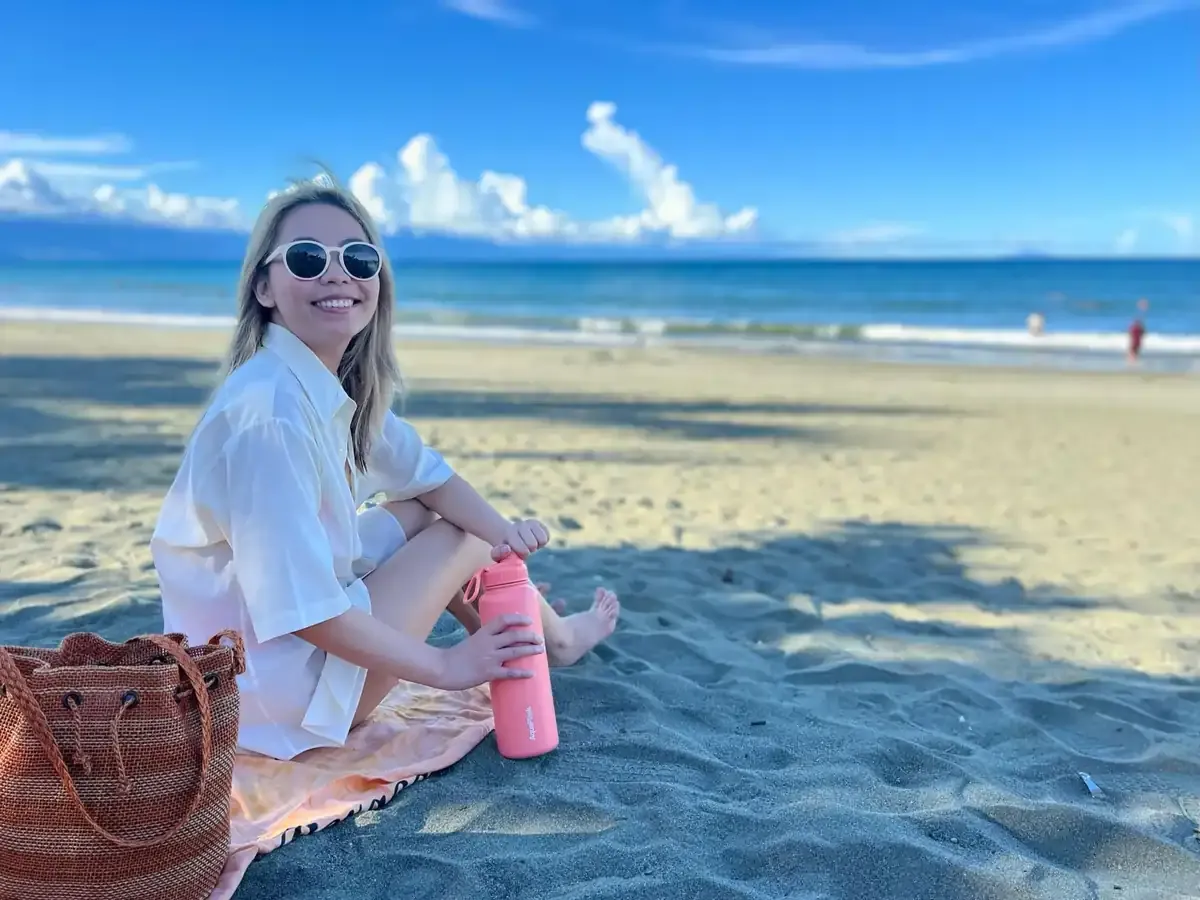 Smiling woman sitting on a beach in the Philippines with a reusable water bottle and beach bag