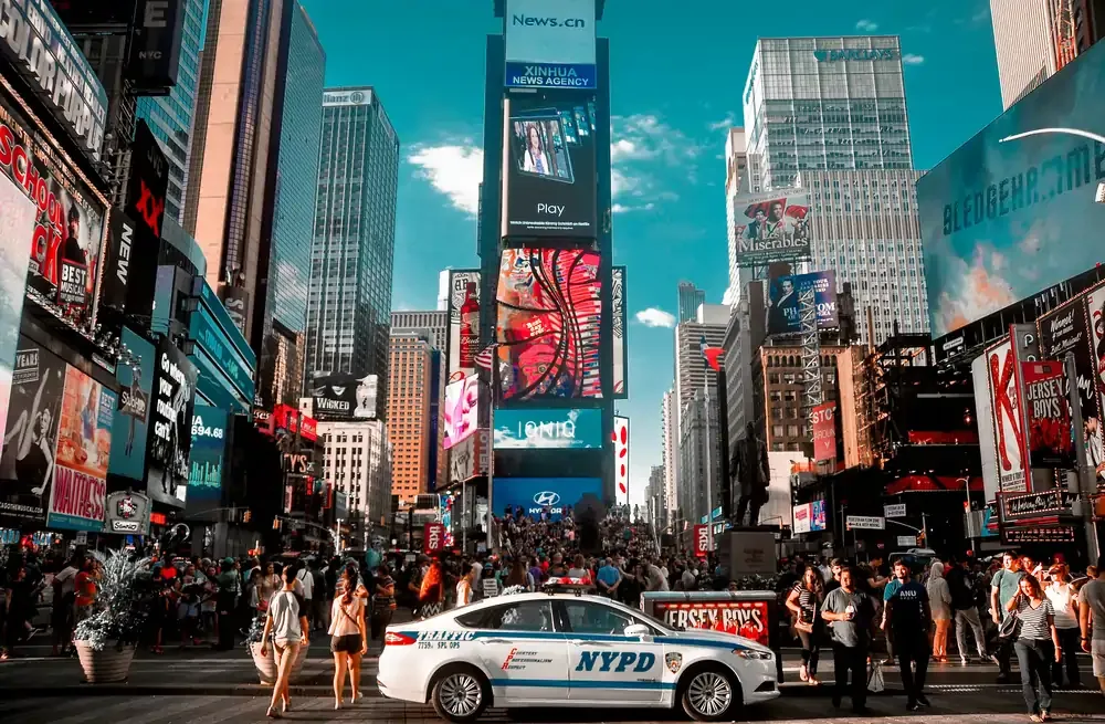 Times Square in New York City with bright digital billboards, crowds of pedestrians, and an NYPD car in the foreground.